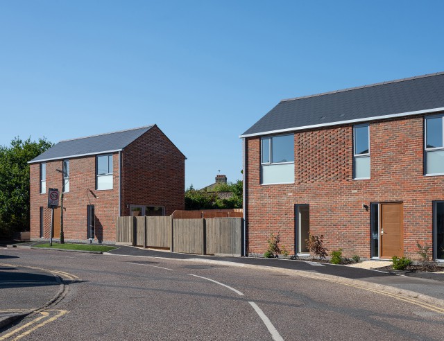 Two red brick modern cottages. Each has tall windows, slate roofs and a section of the wall has  protruding bricks.